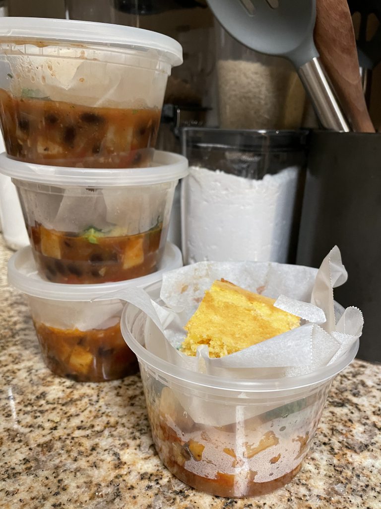 four clear plastic containers filled halfway with chili, then a sheet of wax paper, and a piece of cornbread on top