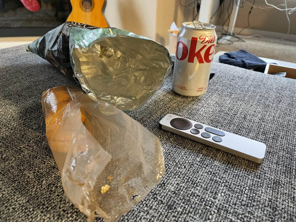 an open cracker sleeve, empty chip bag, Diet Coke can, and an Apple TV remote on a couch cushion